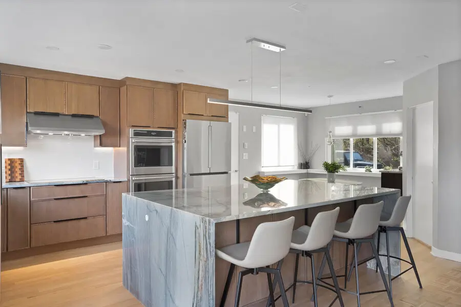 Modern kitchen with a waterfall marble island, warm wood cabinetry, stainless steel appliances, and seating for four.